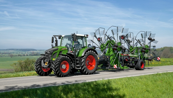 Zielony ciągnik Fendt 600 Vario z czerwonymi obręczami kół jedzie z zieloną zgrabiarką Fendt Former szarą ulicą. W tle widać zieloną łąkę i błękitne niebo.