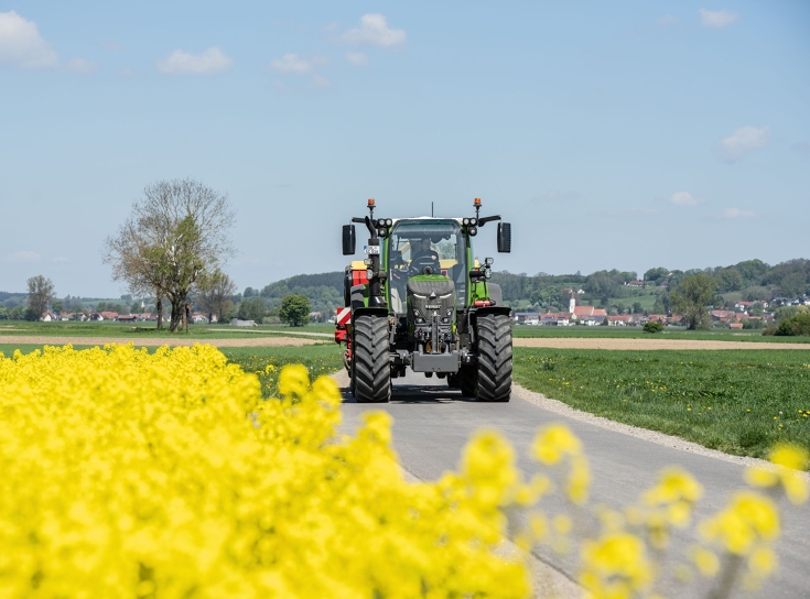 Fendt 500 Vario podczas jazdy na drodze Fendt 500 Vario przejeżdża po drodze, obok pola rzepaku, i transportuje element roboczy z tyłu.