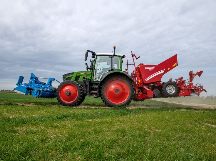 Fendt 500 Vario podczas transportu z sadzarką do ziemniaków i broną talerzową Zielony Fendt 500 Vario z czerwoną sadzarką do ziemniaków z tyłu i niebieską broną talerzową z przodu jedzie po polnej drodze.