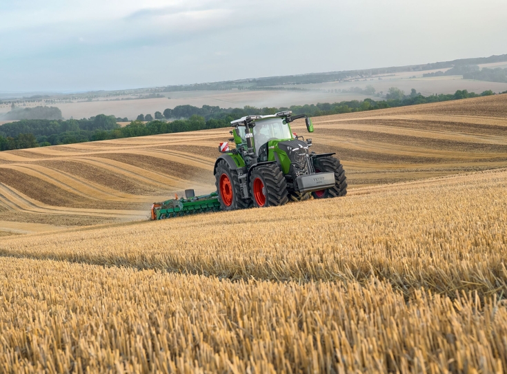 Ciągnik Fendt 800 Vario podczas pracy na polu Zielony ciągnik Fendt 800 Vario jedzie w zestawie z broną talerzową firmy Amazone po ściernisku, w tle widać pola uprawne i las.