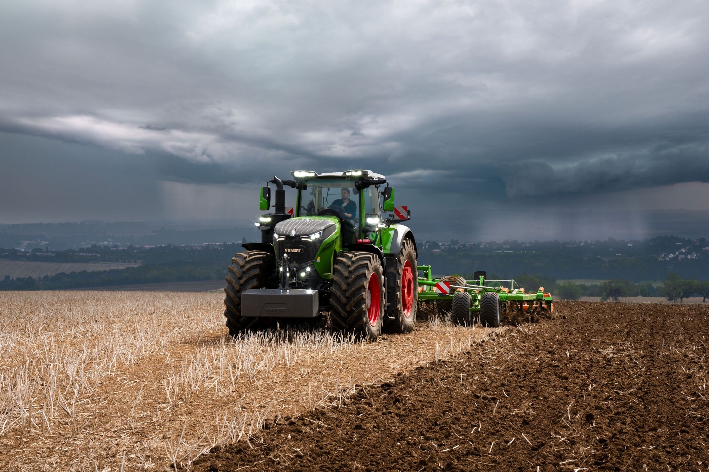 Przed nadciągającymi ciężkimi chmurami burzowymi z oddali ciągnik Fendt 1000 Vario wykonuje prace w polu nadal z pełną mocą