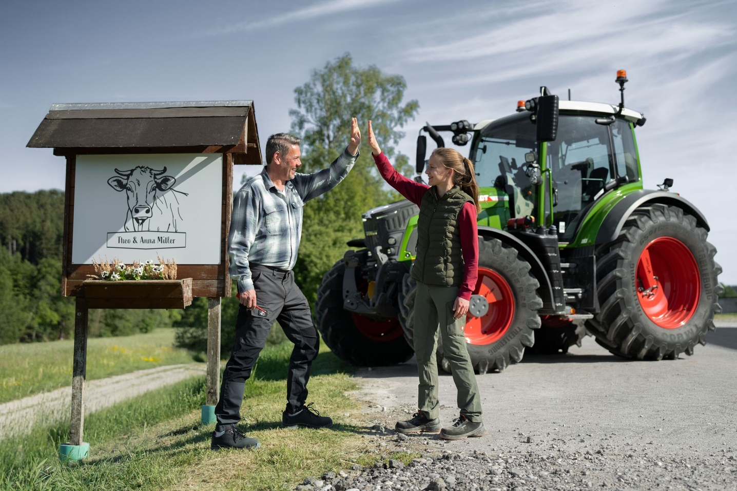 Rolnik i córka stoją przed tablicą gospodarstwa ze swoim Fendt 500 Vario w tle