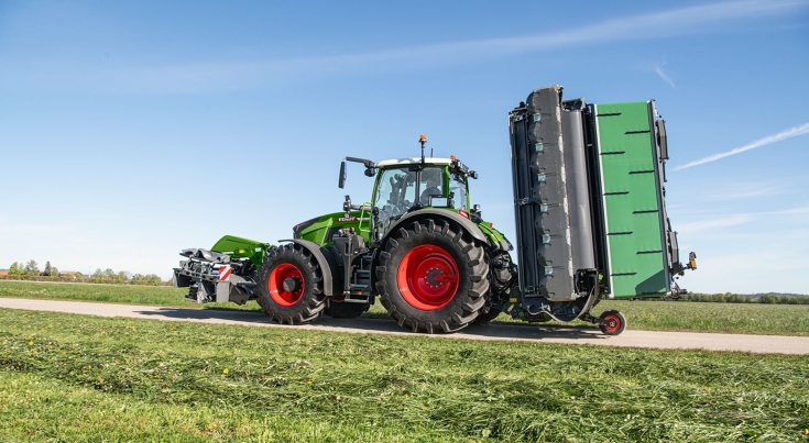 Kosiarka Fendt Slicer z kołem transportowym na drodze Ciągnik Fendt 700 Vario Gen7 porusza się po drodze z kosiarką Fendt mocowaną z przodu i z kosiarką dyskową Fendt Slicer z kołem transportowym.