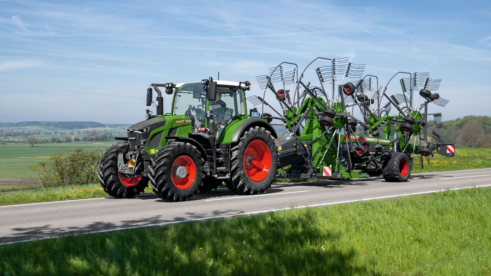 Fendt 600 Vario i Fendt Former Zielony ciągnik Fendt 600 Vario z czerwonymi obręczami kół jedzie z zieloną zgrabiarką Fendt Former szarą ulicą. W tle widać zieloną łąkę i błękitne niebo.