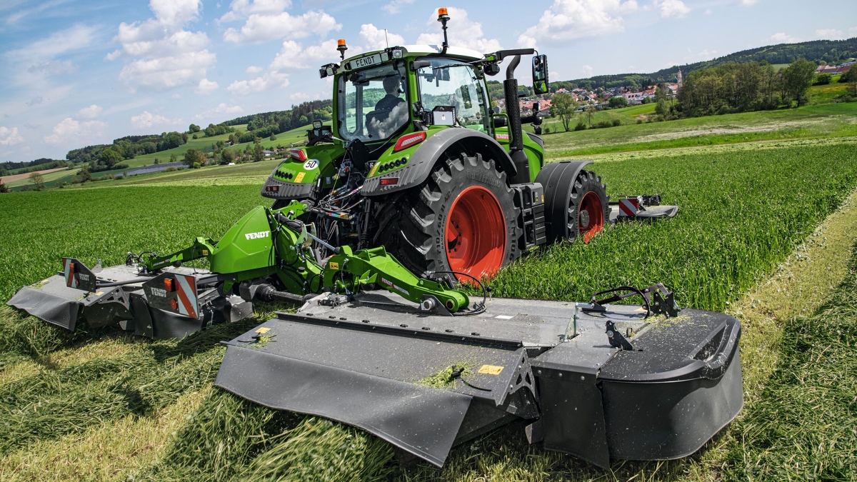 Zielony kombajn Fendt 700 Vario Gen7 jest używany wraz z kombajnem Fendt Slicer na zielonym polu. W tle znajduje się błękitne niebo z białymi chmurami.