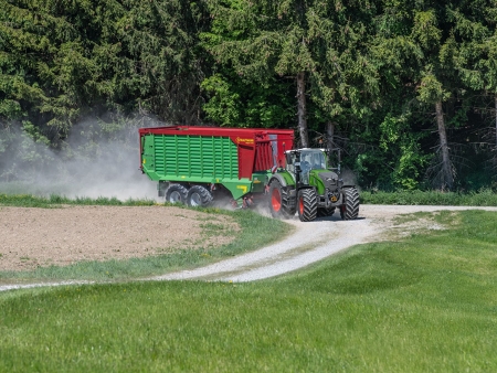 Zielony ciągnik Fendt 724 Vario Gen7.1 jedzie polną drogą z doczepioną przyczepą zbierającą Strautmann. W tle widać las.