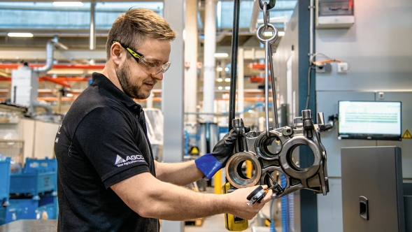 En anställd med AGCO-logotypen på ärmen manövrerar stora maskindelar som hänger från en lyftanordning i en produktionshall. Industrianläggningar och lagerutrymmen syns i bakgrunden.