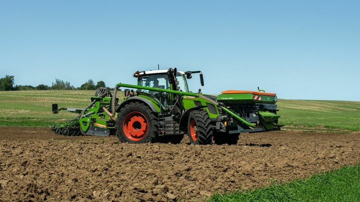 Den gröna Fendt 600 Vario med röda fälgar och en såmaskin som redskapsfäste kör på ett fält. I bakgrunden skymtar en grön äng och en blå himmel.