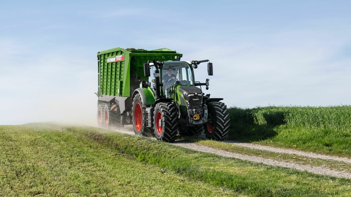 Den gröna Fendt 600 Vario-traktorn med röda fälgar och den gröna Fendt Tigo-självlastarvagnen kör längs en liten åkerväg med en grön äng i förgrunden och bakgrunden.