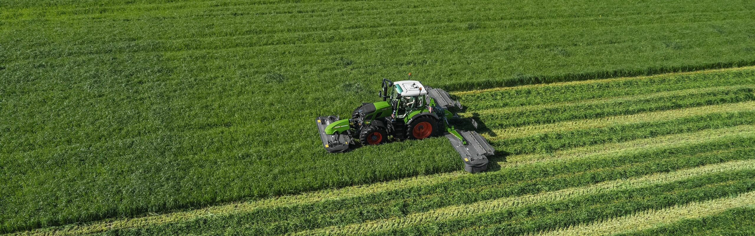 Fågelperspektiv av den gröna Fendt 600 Vario-traktorn i drift med den gröna Fendt Slicer-slåttermaskinen på en stor, grön äng.