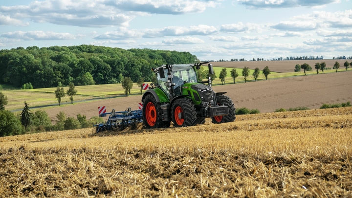 En grön Fendt-traktor med röda fälgar bearbetar jorden i ett skördat fält med böljande kullar i bakgrunden