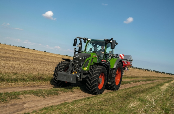 Fendt 600 Vario Den gröna Fendt 600 Vario-traktorn med röda fälgar kör längs en liten landsväg med en ljusblå himmel i bakgrunden.