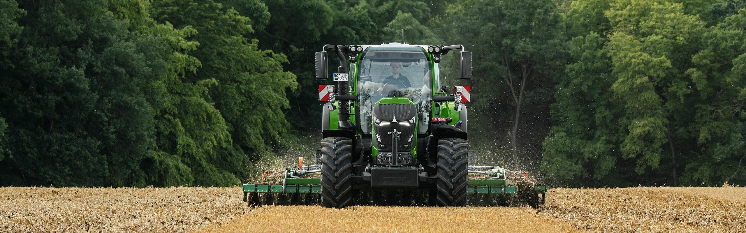 Den gröna Fendt 600 Vario används med ett redskap på en åker. I bakgrunden syns en mörkgrön skog och en molnig himmel.