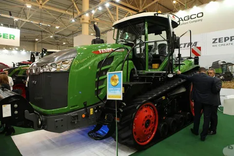 Вигляд спереду зеленого трактора Fendt 1159 MT на гусеничному ходу з червоними колесами, який стоїть у виставковому залі.
