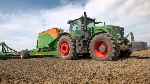 Зелений Fendt 900 Vario з червоними дисками і важким зеленим навісним обладнанням стоїть під хмарним блакитним небом на коричневому полі.