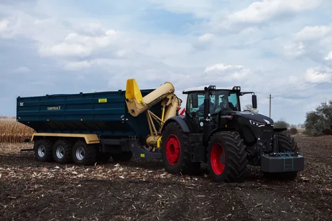 Бічний вид великого чорного трактора Fendt з навантажувальним причепом Egritech, що стоїть на полі із золотими колосками. На задньому плані видно небо з хмарами.