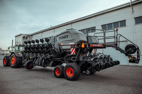 Вигляд ззаду зеленого трактора Fendt з новим Fendt Momentum чорного кольору та сірим бункером для насіння, що стоїть на брукованому дворі перед сірим складом.