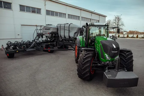 Вигляд спереду зеленого трактора Fendt з новим Fendt Momentum чорного кольору та сірим бункером для насіння, який стоїть на брукованому дворі перед сірим складом.