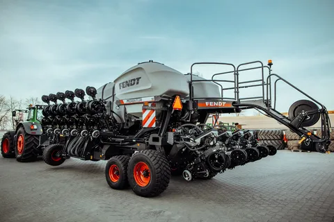 Вигляд ззаду чорного трактора Fendt Momentum з сірими баками та червоними дисками на колесах. На задньому плані — брукований двір фермера, блакитне небо та кілька дерев.