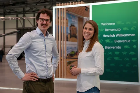 Двоє співробітників компанії Fendt стоять перед зеленою стіною, на якій зображені різні машини Fendt і напис «Ласкаво просимо» різними мовами, і посміхаються в камеру.