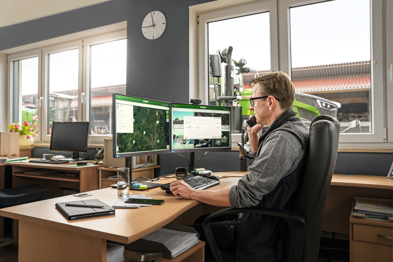 A man sits before of his computer in the office, the documentation of his last tasks are visible on the screens. Through the window, one sees multiple Fendt tractors in a farmyard.