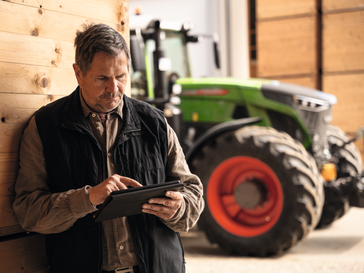 Farm manager stands in a barn next to his tractor and checks his data on a tablet.
