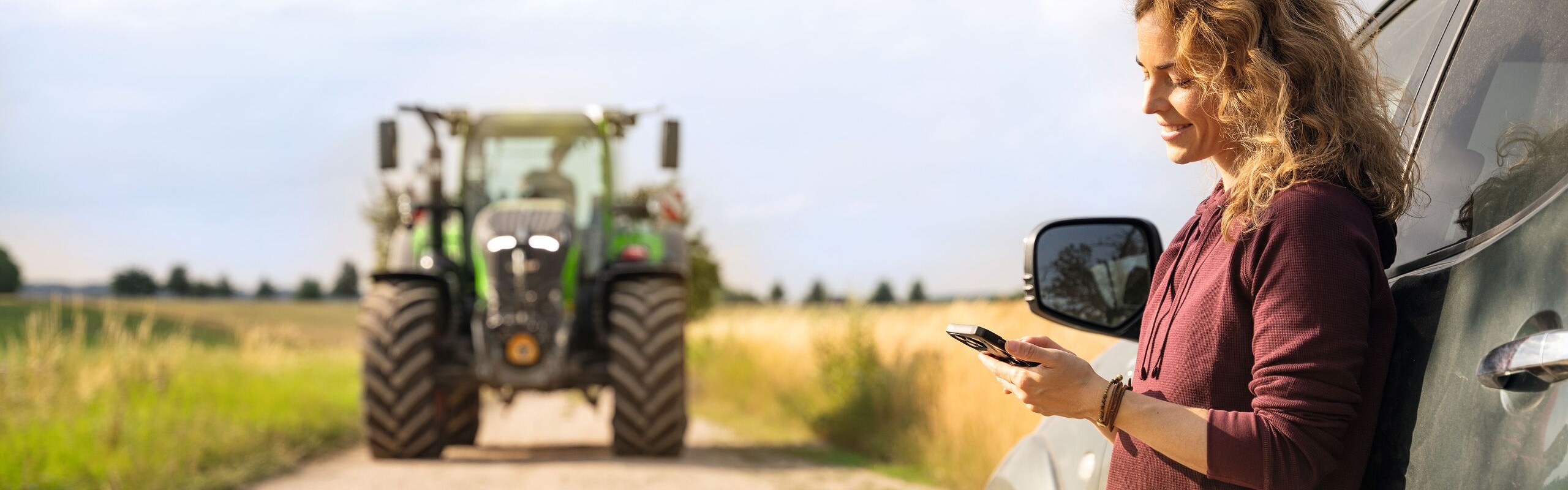A woman with blonde curls and a dark red jumper standing and leaning against a car and looking at her mobile phone; a Fendt tractor can be seen in the background
