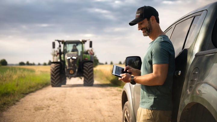 A farmer holding a tablet in his hand and leaning against his car. In the background, a Fendt 700 Vario Gen7 driving up.
