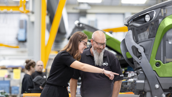 Two employees are standing in a Fendt production hall, looking at a Fendt tractor together. A large green tractor is visible in the foreground, while other work areas, machines, and hall structures can be seen in the background.