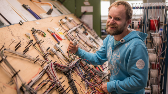 A man in a blue hooded jacket is working on a large wiring board with numerous cables, plug connections and tools. Documents are attached at the top left.