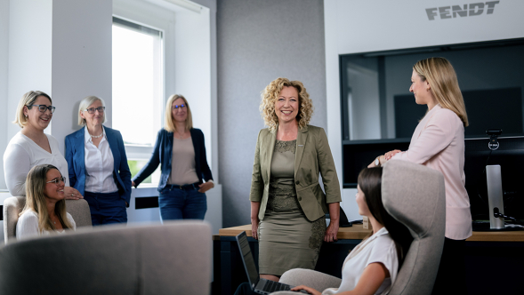 Six women in business attire are in a modern office with a large window and computer desks. The FENDT logo is visible on the wall in the background.