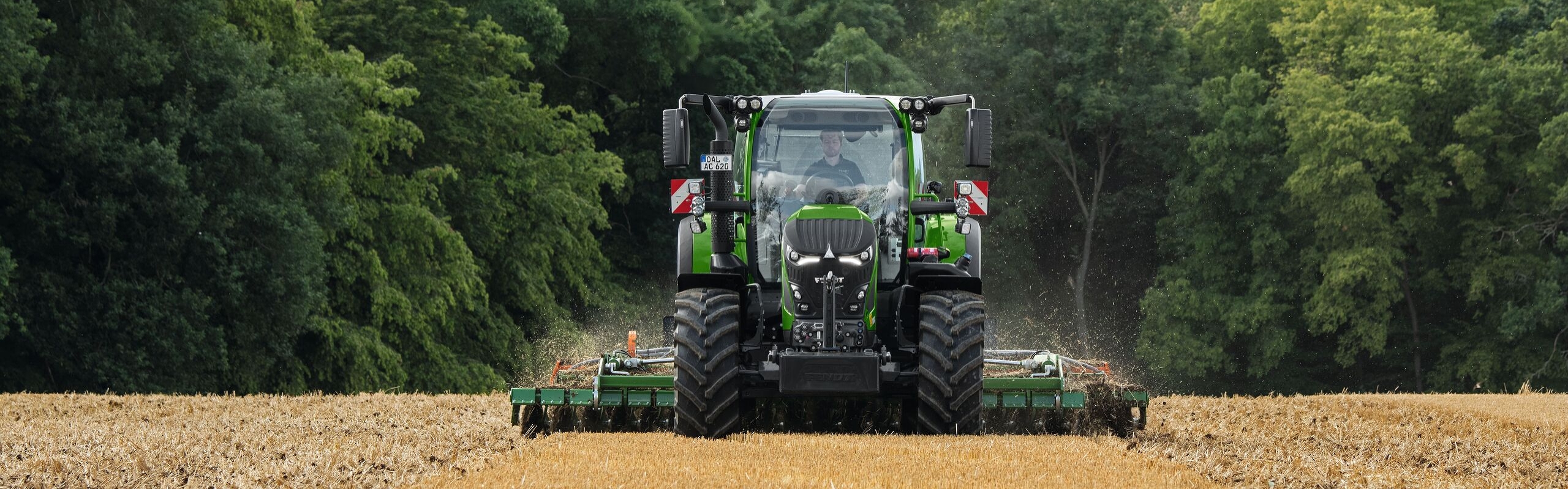 The green Fendt 600 Vario is in use with an implement in a brown field. In the background is a dark green forest and a cloudy sky.