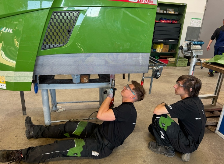 Two men (apprentices) are working on a tractor. One of them is sitting under the bonnet with a hand drill, while the other is crouching down and watching