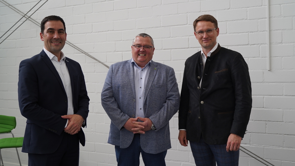 Three men smile together at the camera and stand in front of a white wall