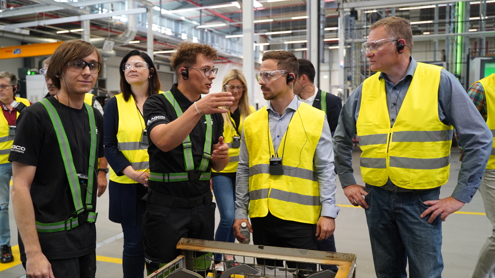 People are standing in a production hall wearing yellow reflective safety vests while one of the two apprentices explains something to them.