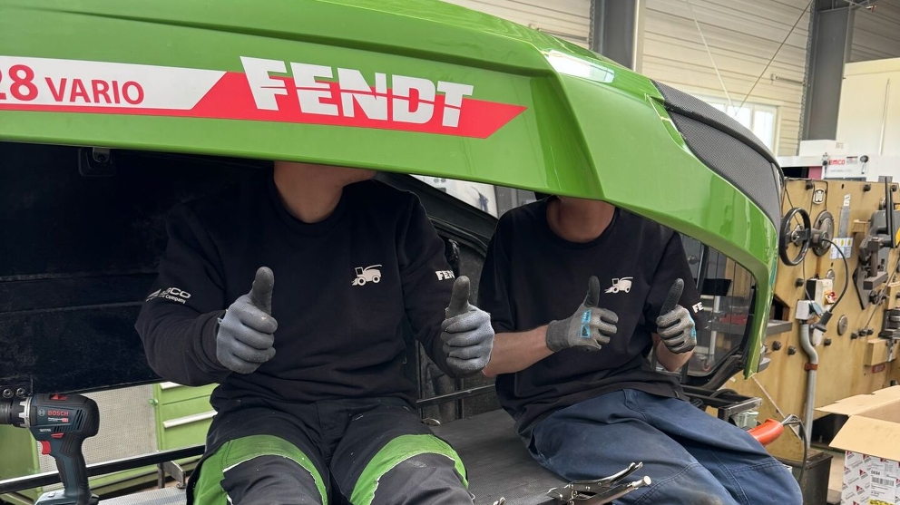2 men (apprentices) sit under the Fendt bonnet and give a thumbs up