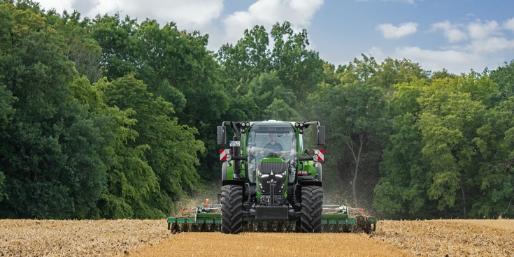 The green Fendt 600 Vario is in use with an implement in a brown field. In the background is a dark green forest and a cloudy sky.
