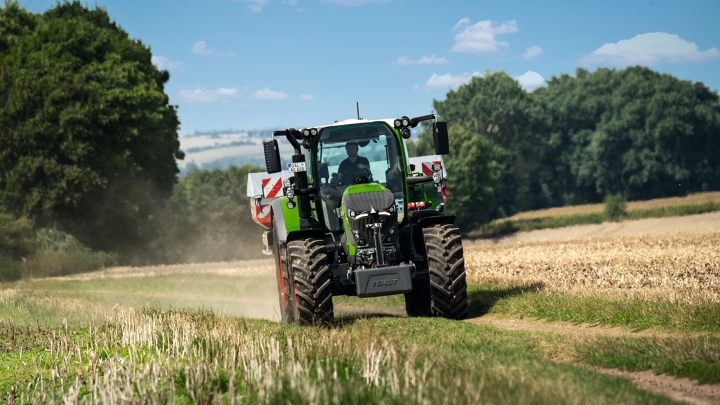 The green Fendt 600 Vario tractor with red rims drives along a small country road with a green meadow in the foreground and a dark green forest and light blue sky in the background.