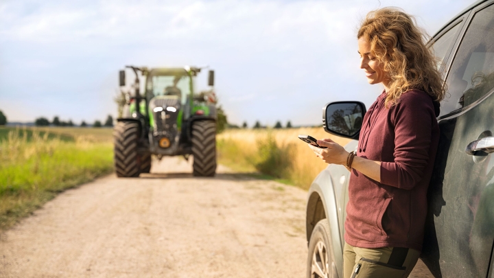 A woman with blonde curls and a dark red jumper standing and leaning against a car and looking at her mobile phone; a Fendt tractor can be seen in the background