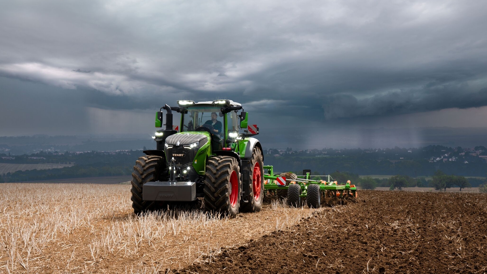 The Fendt 1000 Vario tractor is still doing field work at full power against heavy approaching storm clouds.
