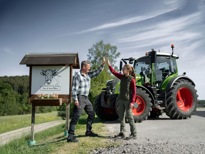 A farmer and his daughter stand in front of their farm sign with their Fendt 500 Vario in the background.