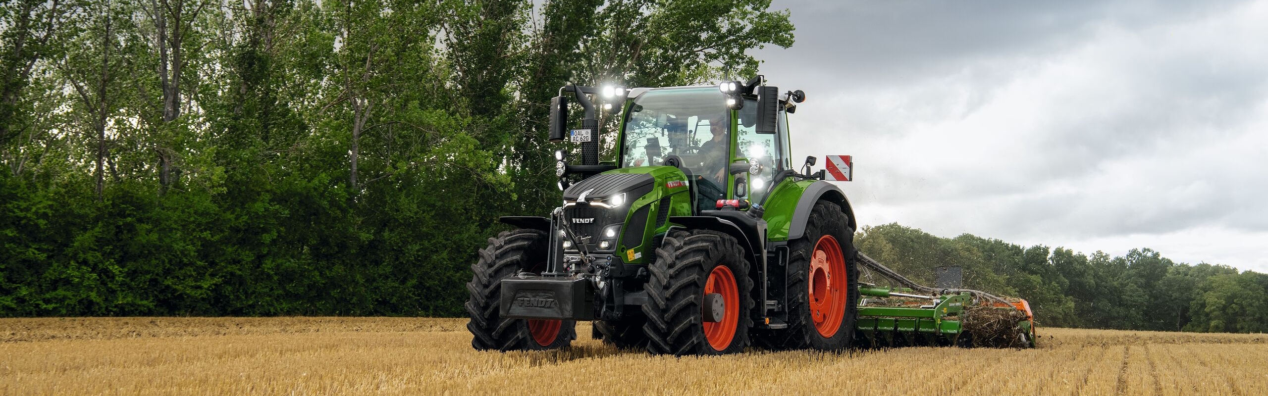 Der grüne Fendt 600 Vario Traktor mit roten Felgen im Einsatz auf einem Acker mit Bäumen im HIntergrund und einem wolkenbehangenen Himmel.