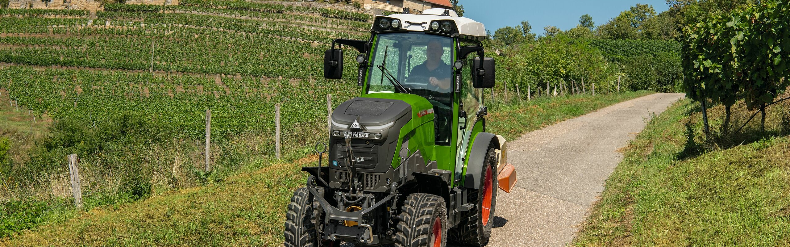The Fendt e100 V Vario A green Fendt e100 V Vario driving between vines on a narrow road.