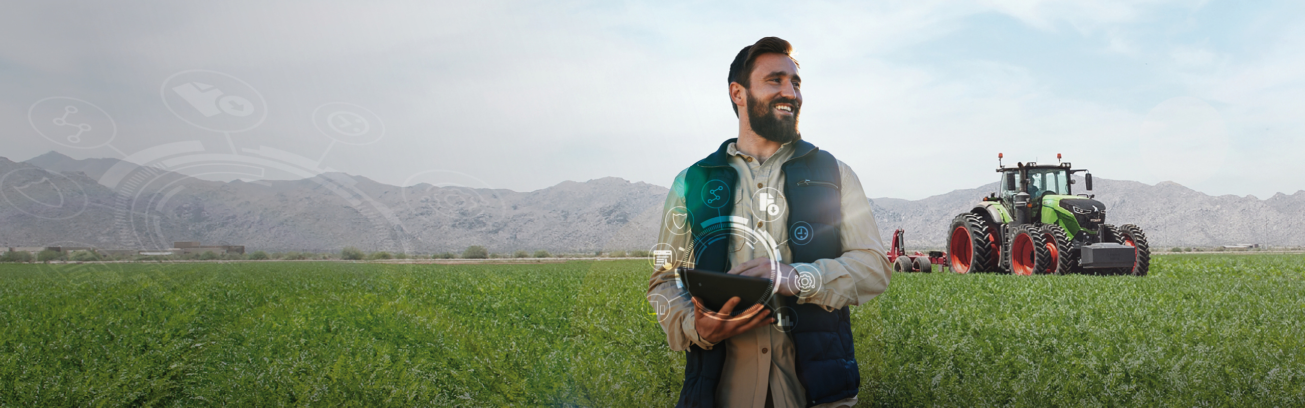 A farmer wtih a tablet stands in a field with a Fendt tractor  and mountains in the background