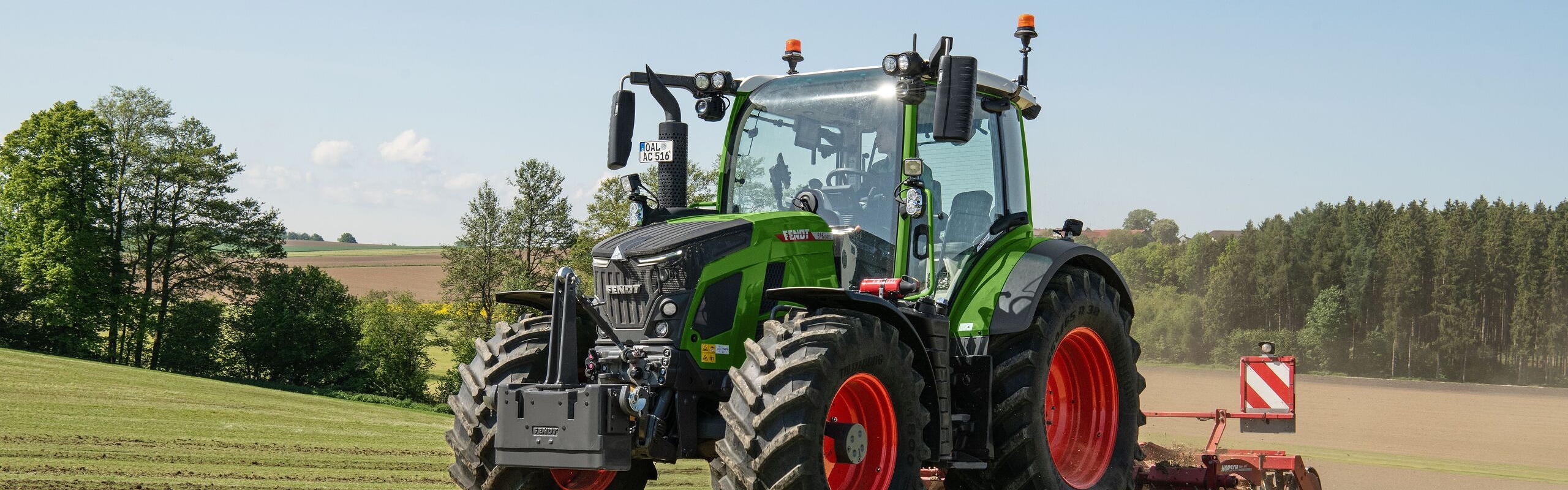 A Fendt 500 Vario Gen4 tractor in green with red rims rides on a light green meadow. At the back is a red soil cultivator with a warning sign attached. In the background are fields, a forest edge and blue sky with few clouds.