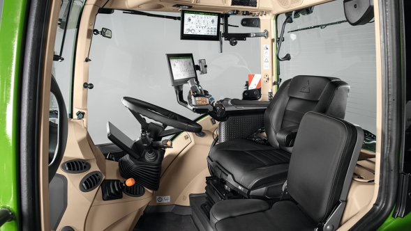 View into the cab of a Fendt 500 Vario Gen4 tractor with beige interior. In the foreground, two black seats – an ergonomic driver's seat and an upholstered passenger seat. On the left is the black steering wheel, next to it an orange control button. On the right side of the cab are digital displays, armrests and control console. The windows let in a lot of light and convey a spacious atmosphere.