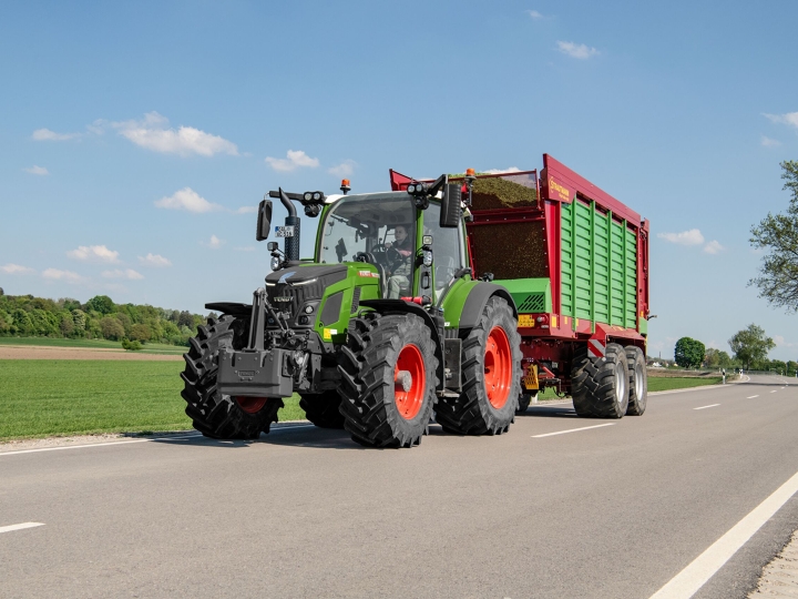 Green Fendt 500 Vario Gen4 with red rims and red forage wagon riding on a grey country road in the sunshine, surrounded by a green meadow and blue sky with white clouds.