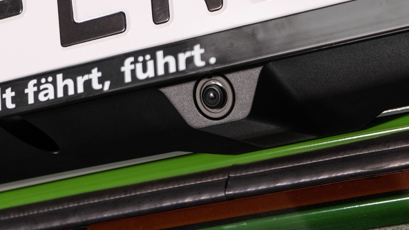 Close-up of a rear camera integrated into the black number plate holder on a Fendt 500 Vario Gen4 tractor. Above the camera is a white license plate with black writing, underneath the green varnish of the body shines.