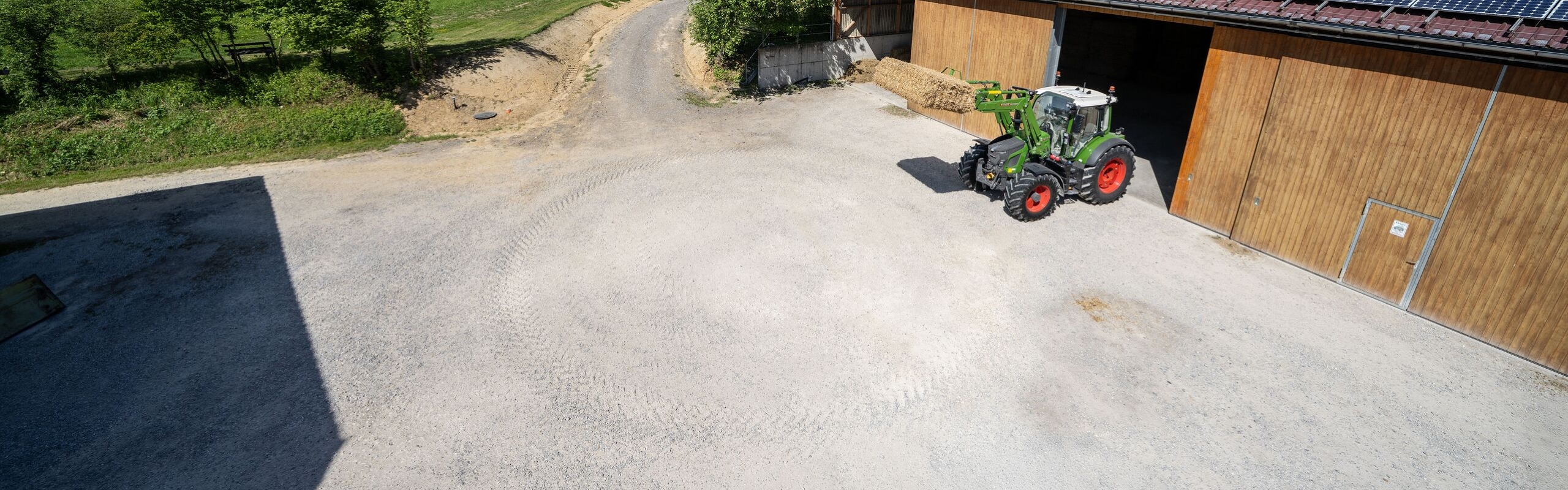 Wide-angle mount of an agricultural farm with a Fendt 500 Vario Gen4 tractor with front loader, transporting a round bale. The courtyard is surrounded by open wooden buildings with solar panels on the roof. The foreground shows a large, bright gravel surface. In the background, a green meadow landscape extends under a blue sky with white clouds.