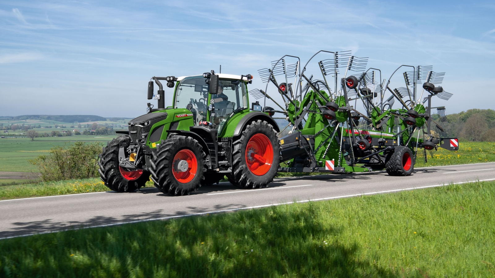 The green Fendt 600 Vario tractor with red rims drives along a grey road with the green Fendt Former. In the background is a large green meadow and a blue sky.